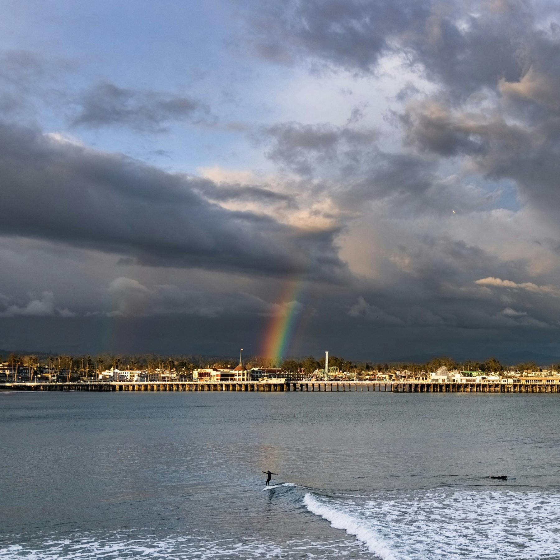 A surfer rides a wave from the nose of his surfboard with the Santa Cruz Municipal Wharf and Beach Boardwalk in the background with a rainbow shining through some dark clouds above captured by Ryan "Chachi" Craig