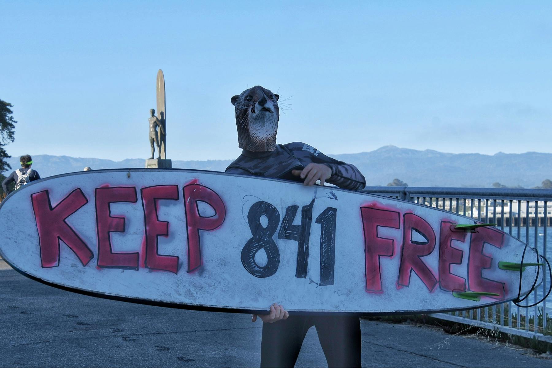 A surfer wearing an otter mask holds up a surfboard the reads 'Keep 841 Free'