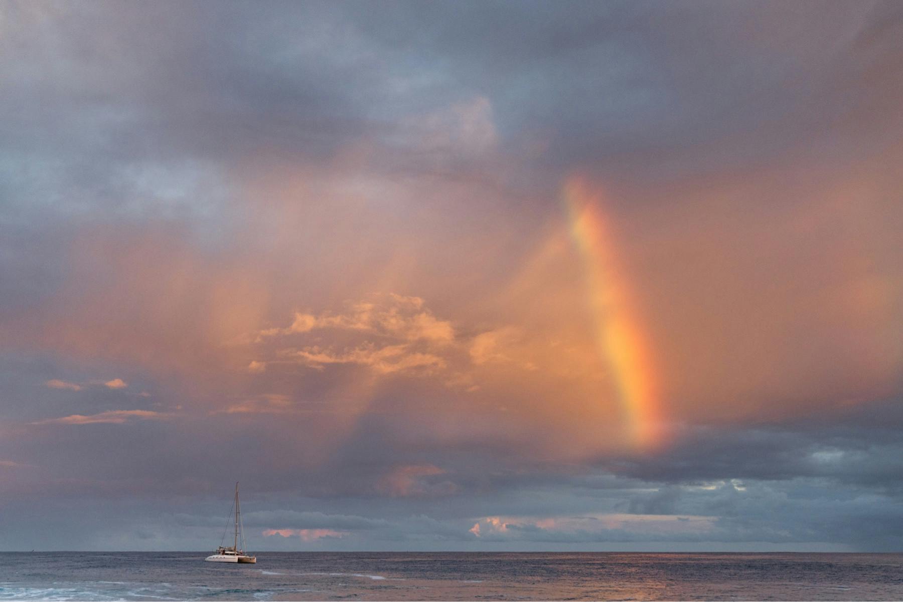 A rainbow beams through the sky at sunrise with a sailboat resting atop the ocean photo by Ryan Chachi Craig