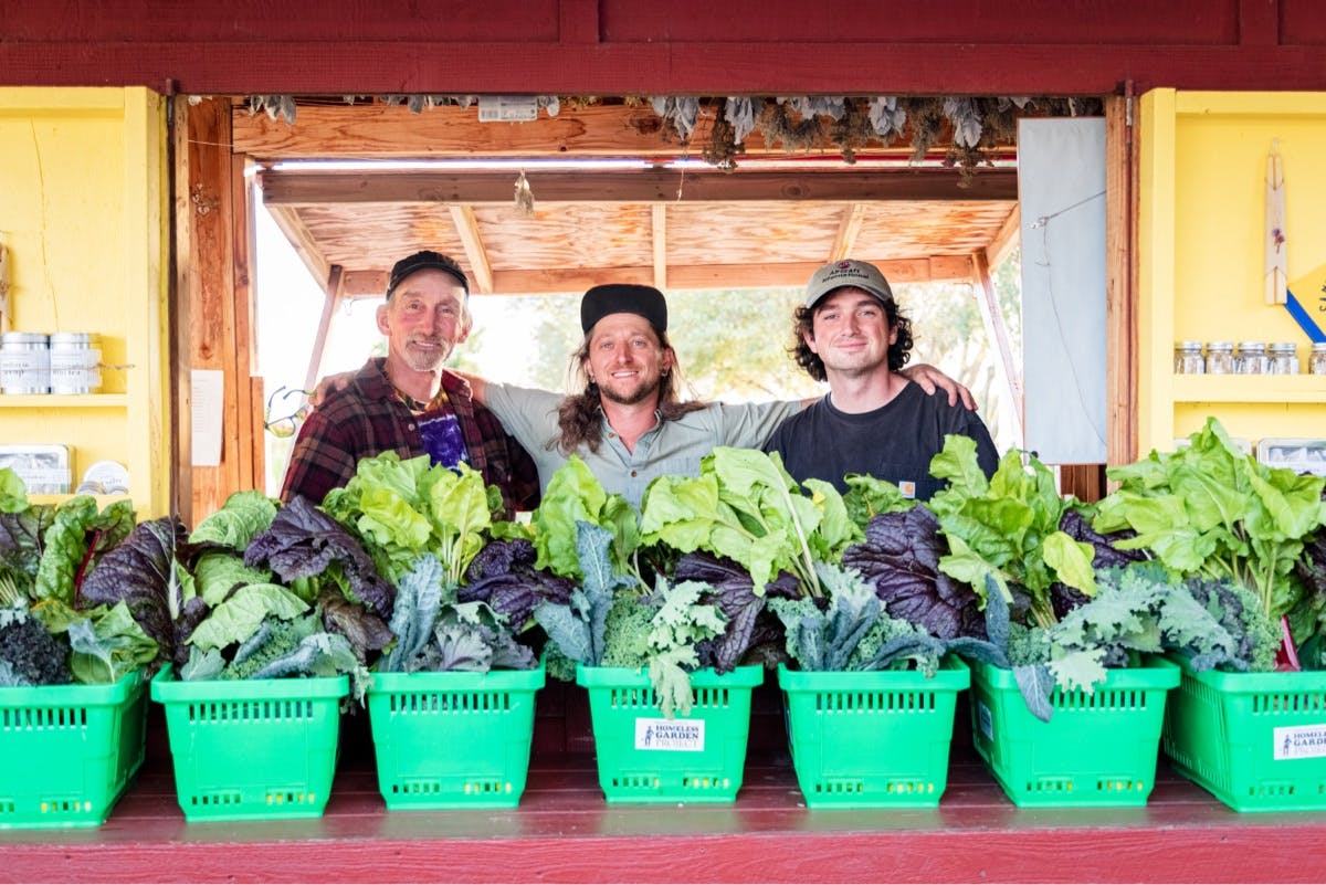 Three attendees of the Santa Cruz Homeless Garden Project stand together in front of freshly harvested produce