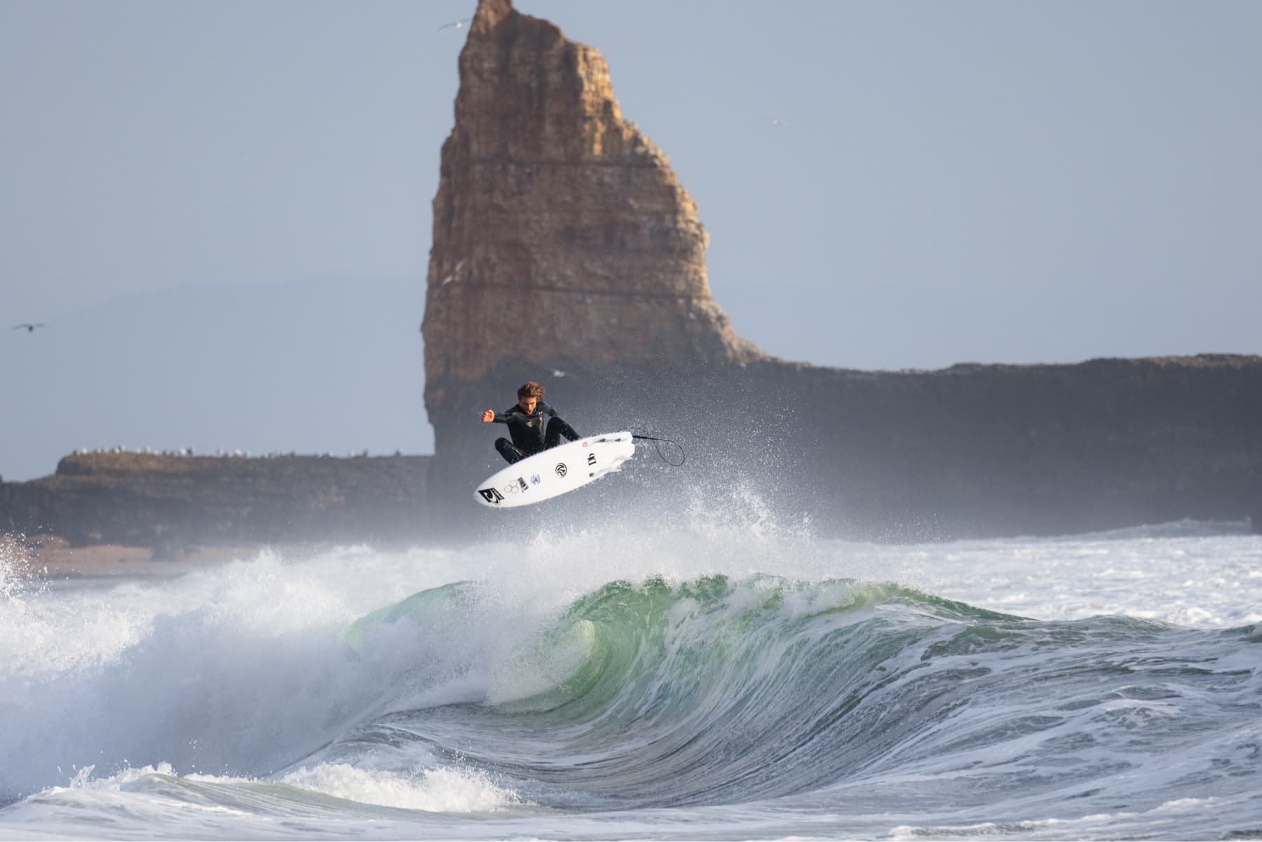 Surfer Noah Wegrich catches air of the lip of a wave with a rock formation in the background captured by Ryan "Chachi" Craig