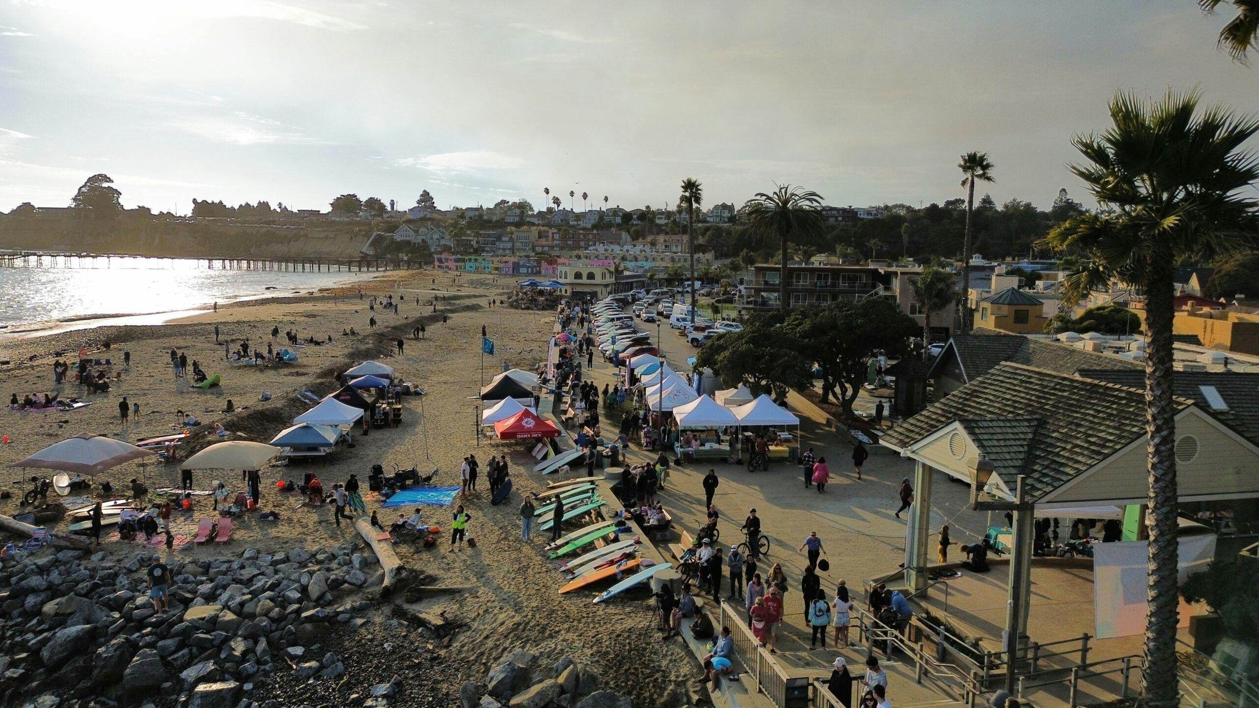 An aerial view of the Capitola Village Esplanade showing surfboards lined up against the wall bordering the sidewalk and beach and numerous people gathering